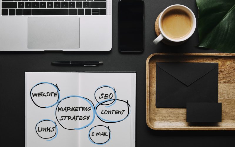 Open notebook with”marketing strategy” icons by stationery and laptop on black background Open notebook with"marketing strategy" icons by stationery and laptop on black background
